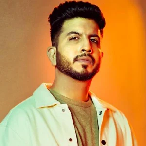 Jigardan Gadhavi, bearded, in a cream jacket against a warm orange backdrop.