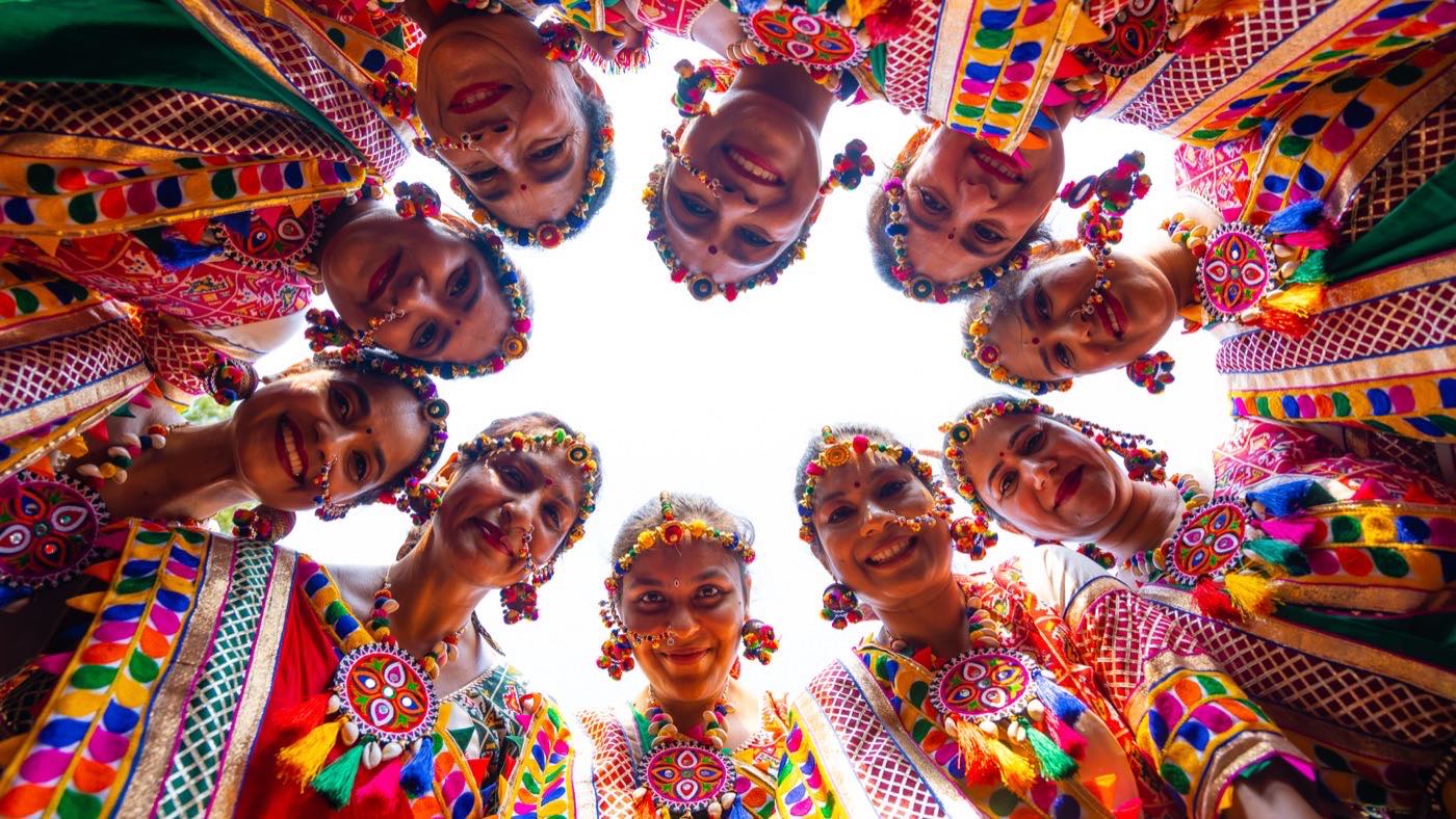 A circle of ten garba dancers in colorful traditional dress, photographed from the center of the circle, all smiling down at the camera