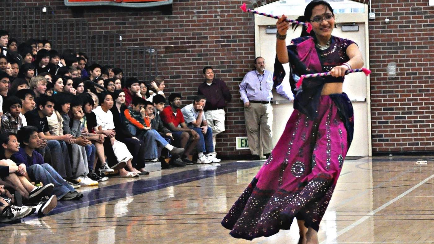 A dancer performing Raas with dandiya sticks in an American school gymnasium, with a crowd watching from the bleachers