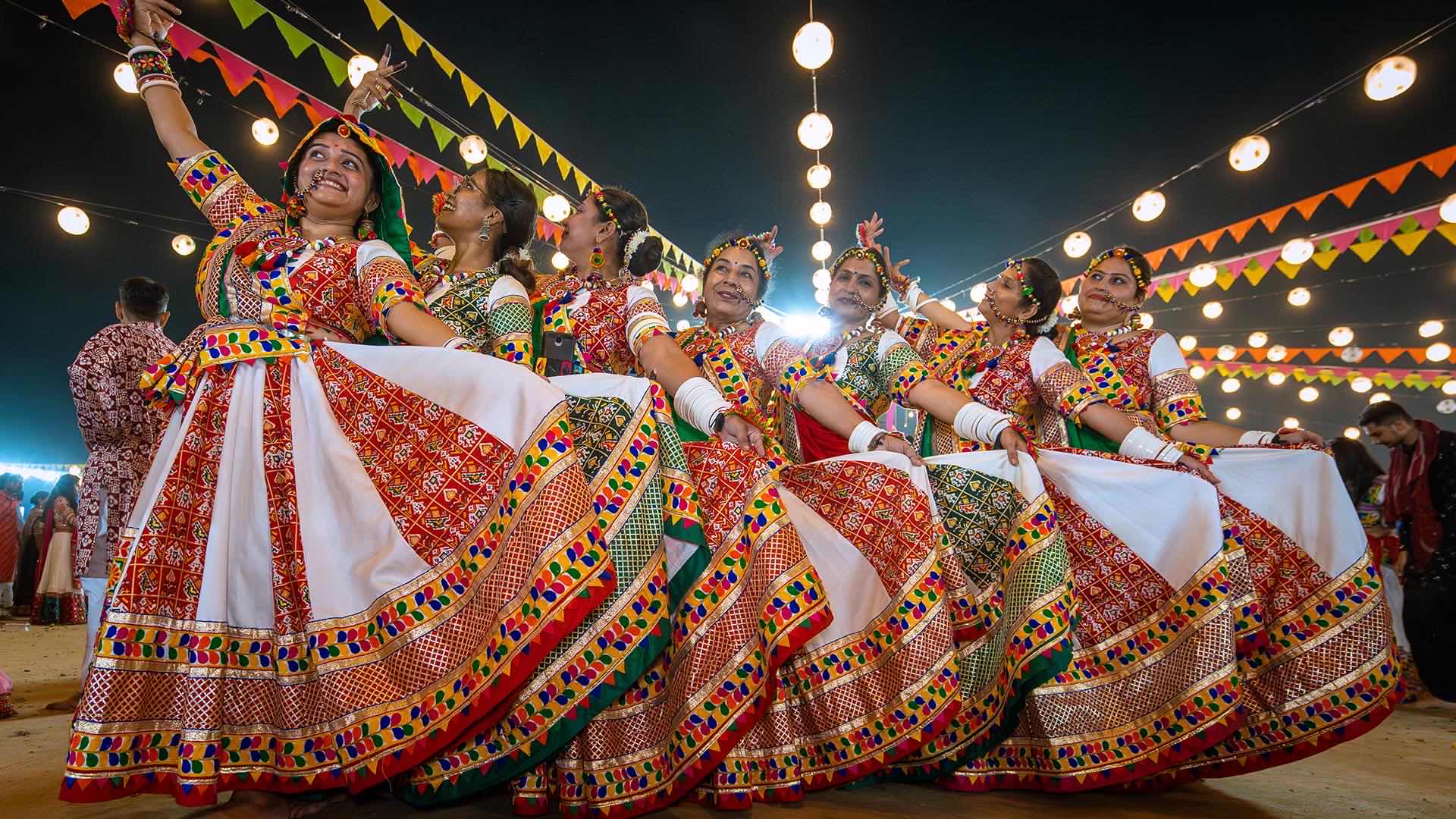 Garba dancers mid-spin in chaniya choli under strings of festival lights at night